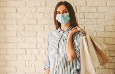 Elinde kağıt torbalarla koruyucu maske takan genç ve mutlu bir kadın. Tıbbi maske takan güzel bir kız alışveriş çantası tutuyor. Kadın alışverişçisi. Uzayı kopyala Coronavirus COVID-19, indirim.