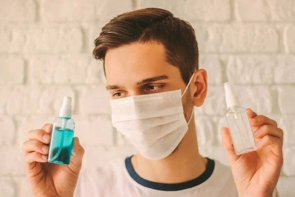 Young man in medical mask on face showing antibacterial gel bottles. Confident doctor in protective face mask holding sanitizers in hands. Personal hygiene, safety. COVID-19 coronavirus protection