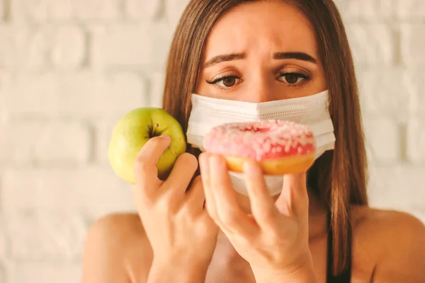 Fitness girl in medical face mask choosing between apple and donut. Sports woman in protective mask deciding between healthy food and sugar snack. Healthy lifestyle, dieting, coronavirus COVID-19