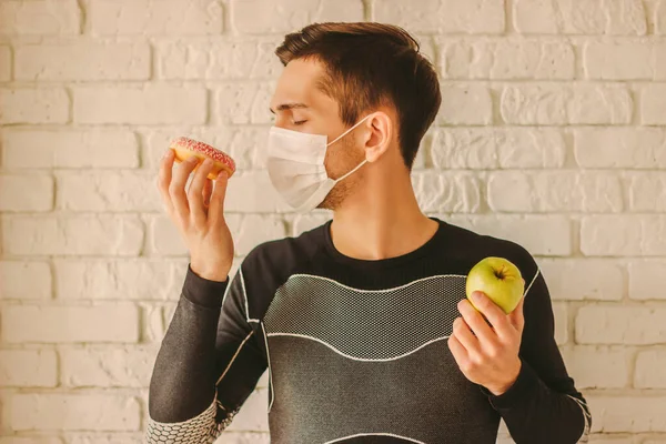 Man fitness trainer in medical face mask holds donut and apple in hands. Sports man in protective face mask choosing between healthy food or sugar snack. Dieting, home quarantine, coronavirus COVID-19