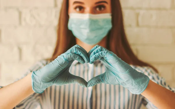 Koruyucu eldivenler ve tıbbi maske takan mutlu kadın kalp sembolü şeklinde el ele tutuşuyor. Tıbbi eldivenli ve koruyucu maskeli genç kız aşk işareti yapıyor. Coronavirus COVID-19, sağlık hizmetleri
