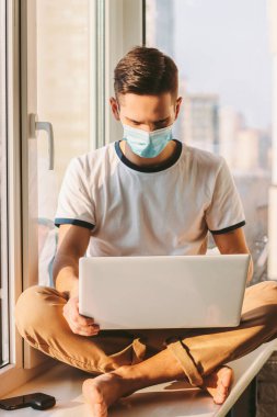 Suratı tıbbi koruyucu maskeli genç adam kucağında dizüstü bilgisayarla pencere eşiğinde oturuyor. Evde çalışan serbest çalışan bir öğrenci. Bilgisayarla mesafeli çalışma. Evden iş. Modern internet günlüğü yazarı