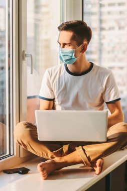Modern internet blogcusu pencere pervazında oturuyor ve dizüstü bilgisayar kullanıyor. Tıbbi koruyucu maskeli genç adam evde çalışıyor. Serbest çalışan öğrenciler için bilgisayarla mesafeli çalışma. Koronavirüs yüzünden karantina