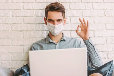 Koruyucu yüz maskeli genç hippi evde video görüşmesi yaparken iyi bir işaret gösteriyor. Tıbbi maskeli mutlu adam kanepeye oturup sanal internet sohbetleri yapıyor. Karantina iletişimi