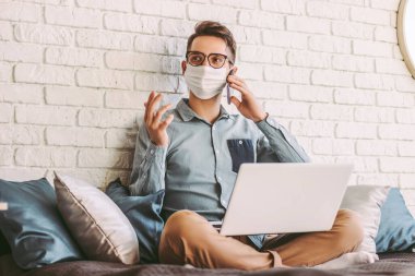 Tıbbi maske takmış, cep telefonuyla konuşan, dizüstü bilgisayarla kanepede oturan mutlu hippi serbest çalışan bir adam. Koruyucu maskeli genç bir işadamı, ofisteki bilgisayarla çalışıyor. Uzak iş