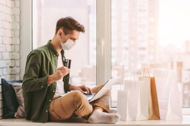 Tıbbi maskeli mutlu hipster evden online alışveriş için dizüstü bilgisayar kullanıyor. Koruyucu maskeli genç adam kredi kartını elinde tutuyor. İnternet alımı için bilgisayar, COVID-19 karantina