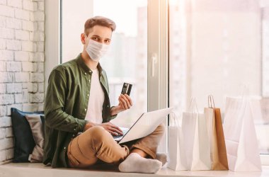 Tıbbi maske takan mutlu adam, internet mağazasından satın almak için kredi kartını elinde tutuyor. Koruyucu maskeli genç hippi evde online alışveriş için dizüstü bilgisayar kullanıyor. COVID-19 karantina