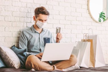 Tıbbi maske takan genç erkek alışverişçi online alışveriş için kanepede dizüstü bilgisayar kullanıyor. Koruyucu maskeli hippi elinde kredi kartı tutuyor. Dijital ödeme. E-ticaret mağazası. COVID-19 ev karantinası
