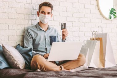 İnternetten alışveriş yapmak için kanepede dizüstü bilgisayar kullanan tıbbi maskeli mutlu adam. Koruyucu maskeli genç hippi elinde kredi kartı tutuyor. İnternet alımları, dijital ödemeler, COVID-19 ev karantinası