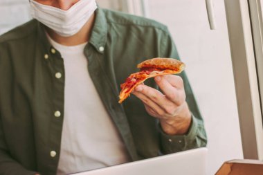 Tıbbî maske takan serbest giyinen, stil sahibi hippi bir adam pizza yer, iş yerinde internette sörf yapar. Aç işadamı elinde pizza dilimi ile dizüstü bilgisayar kullanıyor. Mesafe. İtalyan öğle yemeği molası