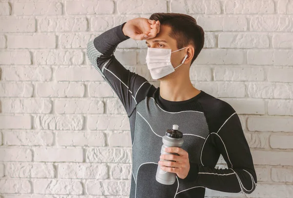 Portrait of tired sports man in medical protective mask on face having rest and drink water after exercising at home. Exhausted fitness man in face mask for personal protection. Quarantine at home