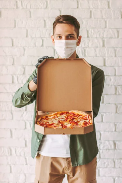 Hipster man courier in protective face mask and gloves carry open pizza box in hands. Happy man delivery service worker in medical face mask hold cardboard box with pizza. Italian fast food delivery