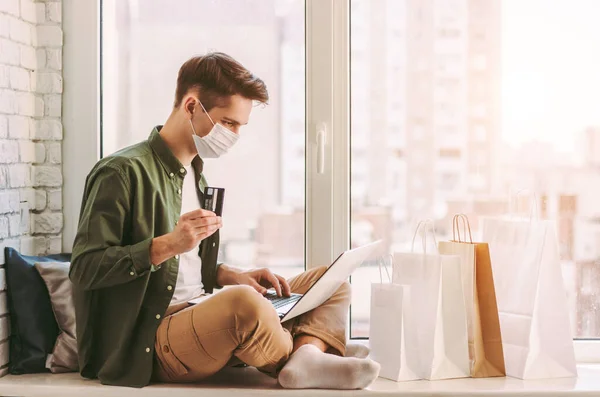 Tıbbi maskeli mutlu hipster evden online alışveriş için dizüstü bilgisayar kullanıyor. Koruyucu maskeli genç adam kredi kartını elinde tutuyor. İnternet alımı için bilgisayar, COVID-19 karantina