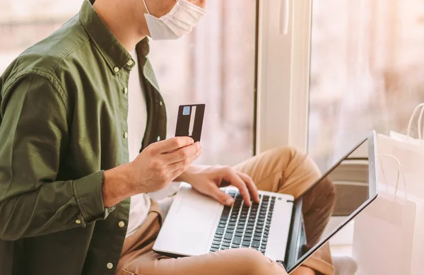 Hipster man in protective face mask hold credit card in hand, typing keyboard, shopping online on laptop. Young man shopper in medical face mask using computer. Internet payment. COVID-19 quarantine