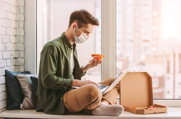 Hungry businessman in medical face mask eat pizza snack, use laptop for work from home office. Stylish hipster man in protective mask hold pizza slice in hand, surf internet, type computer keyboard