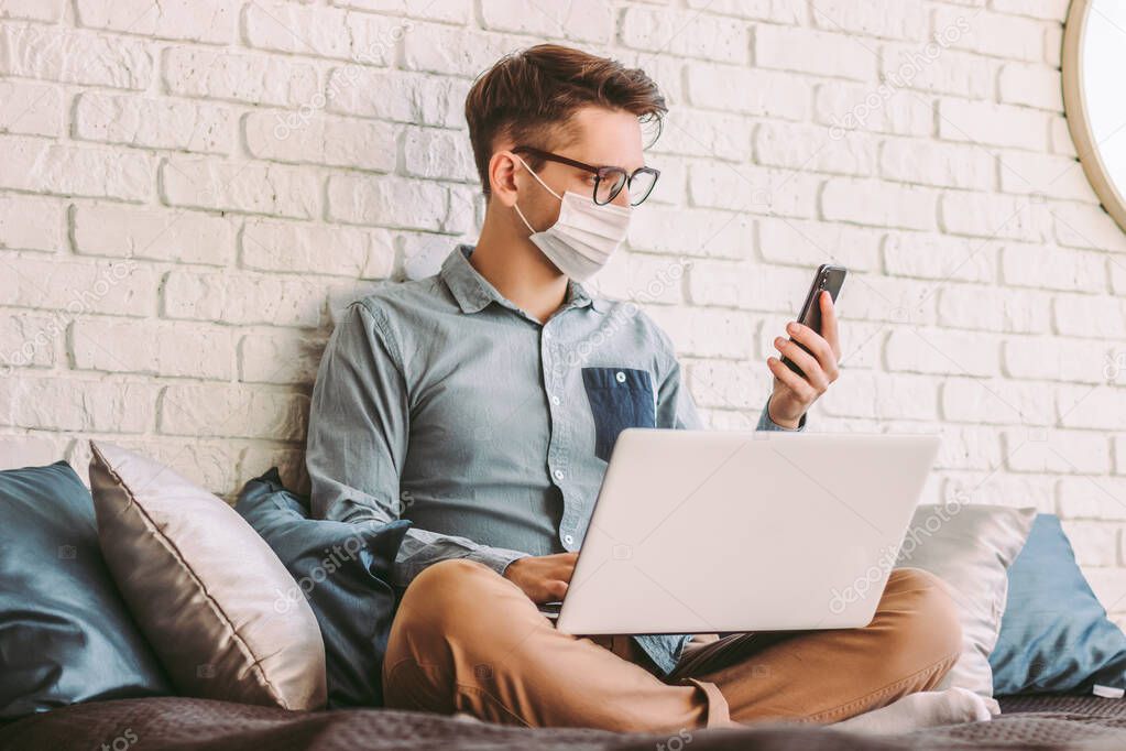 Young businessman in medical face mask using smart phone, texting message while working with laptop at home office. Happy hipster man hold mobile phone in hand, sit on couch. Quarantine communication