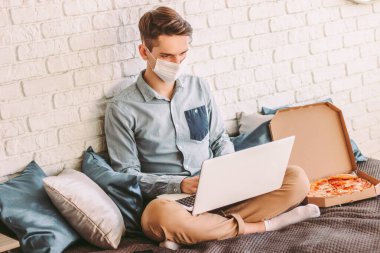 Koruyucu maskeli genç bir iş adamı uzaktan ofis için dizüstü bilgisayar kullanıyor. Medikal yüz maskesi takan serbest çalışan, internetten pizza yiyen, kanepeye oturmakla meşgul bir hippi. COVID-19 karantina