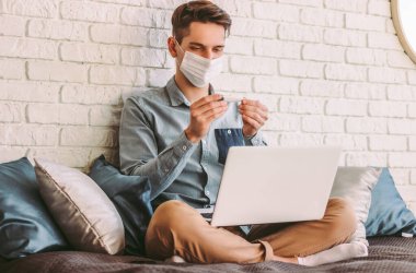 Tıbbi maske takan hasta hippi öğrenci elindeki dijital termometreye bakar, evde çalışır. Koruyucu maskeli genç işadamı elektronik termometre kullanıyor. Vücut ısısını ölç
