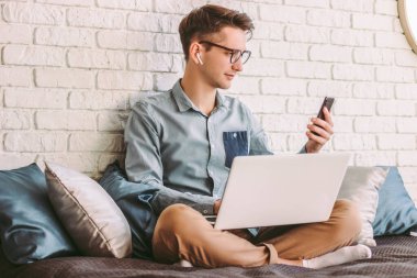 Gözlüklü genç işadamı, cep telefonu kullanıyor, ev ofisinde dizüstü bilgisayarla çalışıyor. Havalı hippi öğrenci akıllı telefondan mesaj atıyor. Çevrimiçi iletişim, uzaktan kumanda, koltukta iş