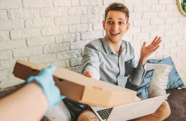 Plastik koruyucu eldivenli POV teslimatçısı mutlu müşteriye karton pizza kutusu veriyor. Hipster adam evde dizüstü bilgisayarla çalışıyor ve hazır yemek alıyor. Güvenli COVID 19 karantina teslimatı