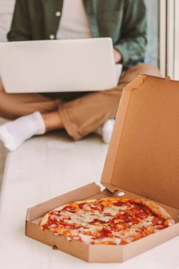 Tahta pencere pervazında taze peynirli pizzayla karton kutuya yakın çekim. Arka planda dizüstü bilgisayarla çalışan hippi bir serbest yazar. Lezzetli İtalyan fast food iş adamları için ev yapımı öğle yemeği