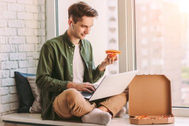 Serbest çalışan genç hippi bir dilim lezzetli pizza yiyor, pencere kenarına oturuyor ve çalışmak için dizüstü bilgisayar kullanıyor, uzaktan öğreniyor. İş adamı öğle yemeği için İtalyan fast food 'u yer, evden uzakta çalışır.