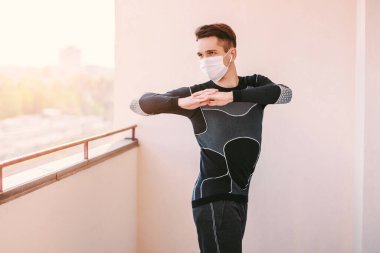 Koruyucu maskeli mutlu genç sporcu COVID-19 ev karantinası sırasında kaslarını germek için yoga egzersizleri yapıyor. Kendine güvenen bir fitness eğitmeni. Balkonda maske eğitimi alıyor.