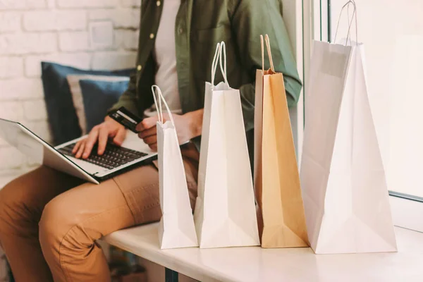 Bilgisayarı online alışveriş için kullanan, kredi kartını elinde tutan, pencere eşiğinde çantalarla oturan genç hippi. Evden internet mağazasından alışveriş yapan şık bir adam. E-ticaret, perakende