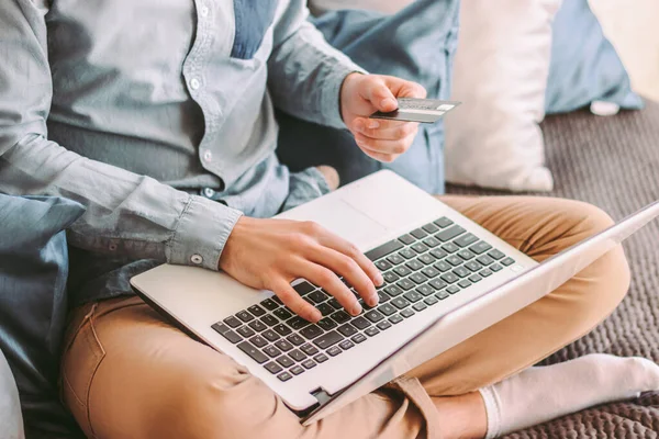 Closeup stylish male shopper holding credit card in hand and typing laptop keyboard, sitting on couch. Young hipster man shopping online with computer at internet store. E-commerce purchase from home