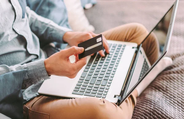 Kapalı kapılar ardında genç hippi erkek müşteri kredi kartı tutuyor ve dizüstü bilgisayarda klavye çalıyor, internetten alışveriş yapıyor, evde çalışıyor. İnternet alışverişi ve ödeme için bilgisayar kullanan şık bir serbest çalışan. E-ticaret