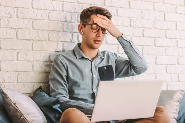 Young stressed hipster man in glasses feel tired, exhausted while use laptop on couch. Nervous frustrated freelance businessman look unhappy or depressed, work at home. Receive bad news, trouble, fail