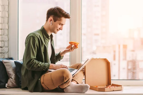 Happy businessman eat slice of tasty pizza, work remotely from home. Young hipster man freelancer sit on windowsill, using laptop for distance education, work, study, eat italian takeaway fast food