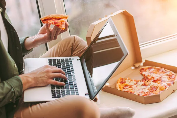 Yakından stil sahibi serbest iş adamı lezzetli bir dilim pizza yer ve evden uzakta çalışmak için dizüstü bilgisayar kullanır. Hippi adam İtalyan fast food atıştırmalıklarını elinde tutuyor, klavyede yazıyor. Yemek molası.