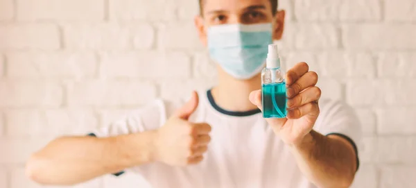 Banner confident doctor in protective mask on face holding sanitizer bottle in hand and showing like thumb up. Young man presenting antibacterial gel as preventive measure against coronavirus COVID-19