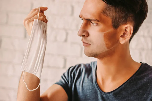 Portrait of angry young man with funny sunburn lines holding medical face mask in hand. Confused hipster man with sun tanned lines on face after wearing protective face mask. COVID-19 prevention