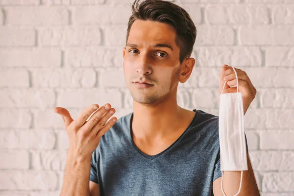 Portrait of confused hipster man with sunburn tanned face skin holding protective mask in hand. Young man patient with sun tanned lines on face after wearing medical face mask. COVID-19 quarantine