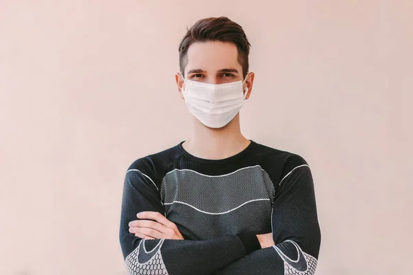 Medikal maskeli sporcu portresi kollarını kavuşturup arka planda gülümsüyor. Genç ve mutlu fitness adamı. Profesyonel kişisel antrenör evde. COVID-19 koronavirüs karantinası