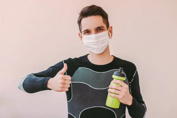 Happy man fitness instructor in medical face mask showing thumb up gesture. Young confident sports man in protective mask gesturing like for training at home during coronavirus COVID-19 quarantine