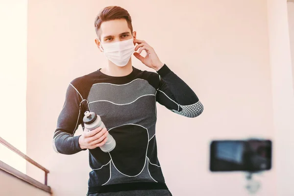 Confident sports man in medical face mask recording video tutorial for training at home during COVID-19 quarantine. Young fitness instructor blogger media influencer training against phone camera