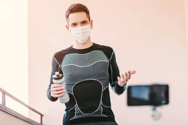 Young man fitness coach in medical face mask training against phone camera. Confident sports man in protective mask record video tutorial for fitness blog. Home training, workout, COVID-19 quarantine