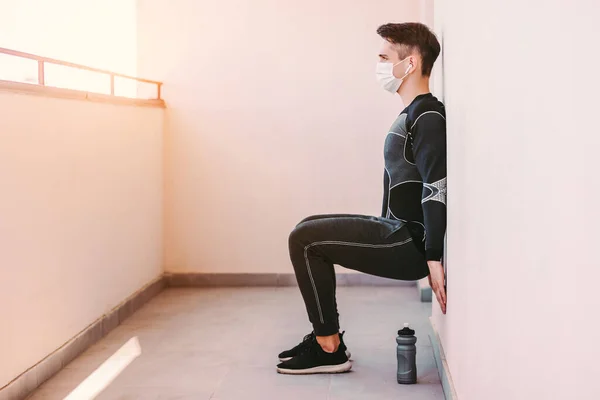 Tıbbi yüz maskesi takan genç sporcu fitness koçu balkonda duvara yaslanıyor. Koruyucu maskeli kendine güvenen sporcu, spor salonunda duvar bacağı egzersizi yapıyor. COVID-19 karantina, çalışma