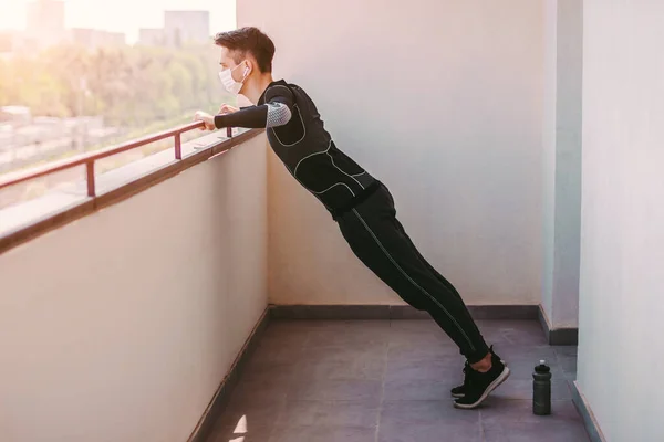 Koruyucu yüz maskeli genç sporcu evde şınav çekiyor. Balkonda tıbbi maske takan sportif kendine güvenen fitness antrenmanı yapan adam. COVID-19 karantina. Ev egzersizleri