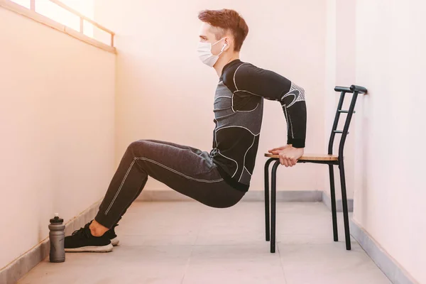 Medikal maskeli genç sporcu evde sandalyede triseps dalışı yapıyor. Koruyucu maske takan sportif atletik erkek fitness eğitmeni triseps kası için egzersiz yapıyor. COVID-19 karantina