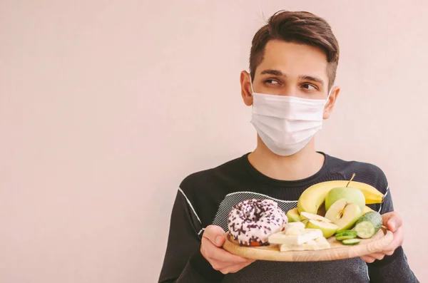 Uncertain sports man in medical face mask hold wooden board with junk and healthy food, look at copy space. Fitness man in protective mask struggling: sweets or fruits. Quarantine dieting nutrition