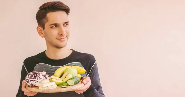 Hungry young sports man holding wooden board with fresh fruits, vegetables, sweet donut, chocolate and look at copy space. Happy fitness man choosing between healthy food and junk unhealthy snacks