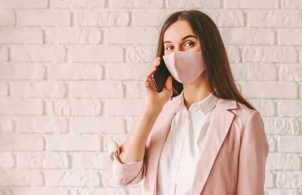 Portrait professional young business woman in stylish suit, medical face mask talk cell phone and smile. Happy beautiful girl entrepreneur in pink protective face mask with smart phone. Communication