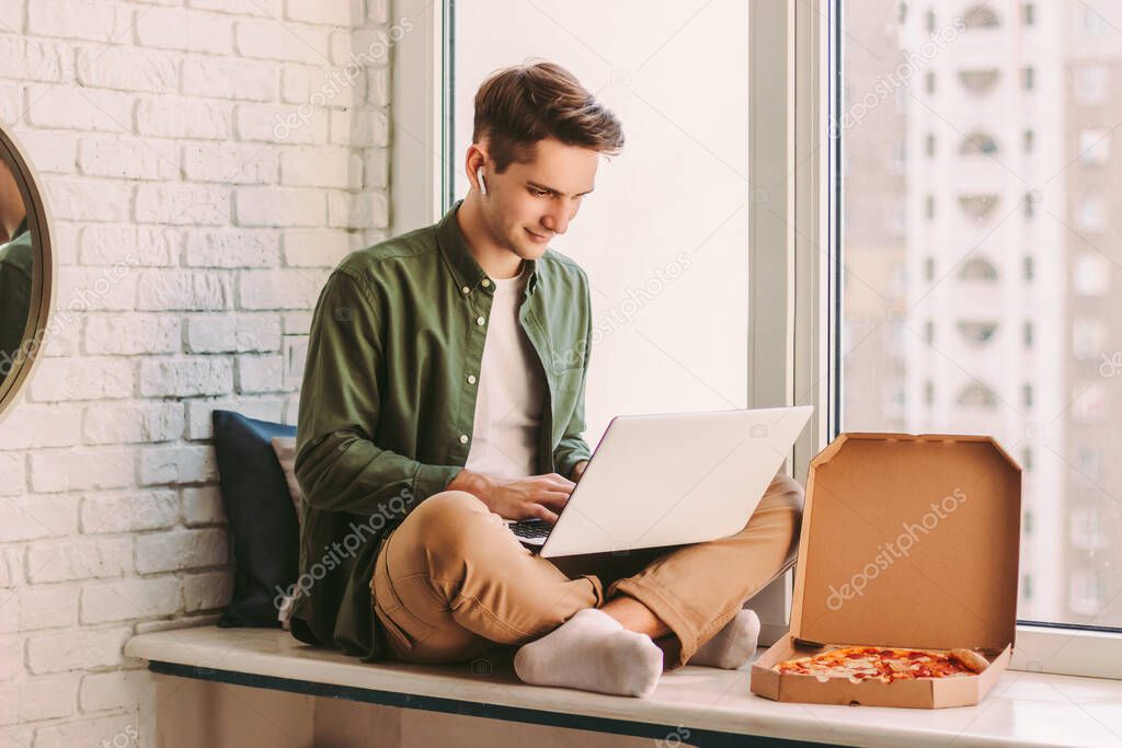 Happy businessman using laptop for distance work from home and eat tasty takeaway pizza fast food. Young hipster man freelancer typing on computer keyboard, study, remote education, sit on windowsill