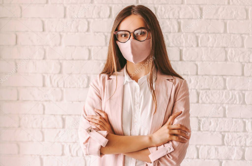 Portrait confident young female entrepreneur in protective face mask, suit and eyeglasses keep arms crossed. Beautiful professional business woman in glasses wearing pink medical face mask. Girl boss