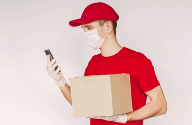 Portre teslimatçısı üniformalı, tıbbi maske takan, koruyucu eldiven takan cep telefonu kullanan, arka planda kargo kutusunu elinde tutan. Teslimat işçisi sevkiyat ve akıllı telefon taşıyor.