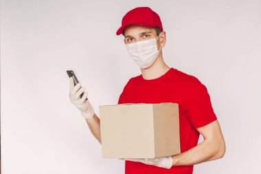Tıbbi maske takan mutlu teslimat servisi kuryesi koruyucu eldivenler beyaz arka planda izole edilmiş akıllı telefon ve karton kutular tutuyor. Paket kutusu olan genç teslimatçı cep telefonu kullanıyor.